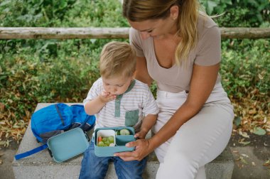 Günlük kıyafetlerle gezen anne, anaokulundaki küçük çocuğun yanında oturuyor ve güneşli bir günde okuldan sonra birlikte vakit geçirirken öğle yemeğinde taze sebze yiyor.
