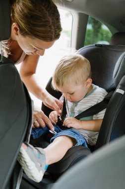 A young smiling mother fastens seat belt on the child seat in which her little child is sitting. the concept of safe trip and health insurance.