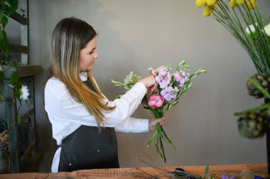 Side view of young female florist in apron arranging bouquet of various flowers while standing by table and working in floral shop