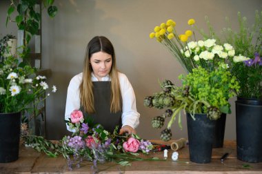 Young focused female florist in apron standing with freshly cut aromatic flowers and blooming potted flowers on table while working in floral shop. business content