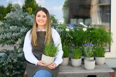 Positive young female florist and business owner in casual clothes with apron smiling and looking at camera while sitting on bench with green potted blooming flowers. business content