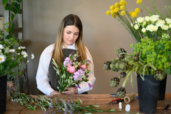 Young female florist standing at table with bouquet of aromatic flowers and potted flowers in green leaves while working in floral shop