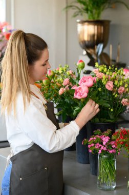 Çiçekçide çalışırken sarı saçlı, pembe gülleri vazolara yerleştiren genç bayan çiçekçinin yan görüntüsü.