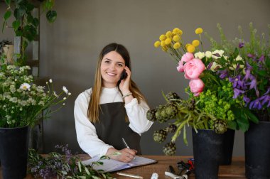 Önlüklü, gülümseyen, akıllı telefonlu genç bir çiçekçi. Dükkandaki çeşitli çiçeklerin yanında deftere bir şeyler yazarken.