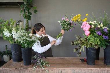 Önlüklü kadın elinde çiçeklerle ahşap masada duruyor ve çiçekçi dükkanındaki kökleri kesiyor.