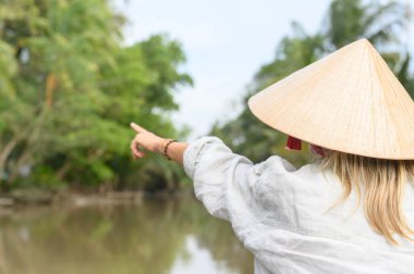 Vietnam 'da Mekong Deltası' nda turne sırasında ağaçlara işaret eden günlük giysiler içinde Vietnamlı bir şapka takan Kafkas genç sarışın turistin arka görüntüsü. Yüksek kalite fotoğraf