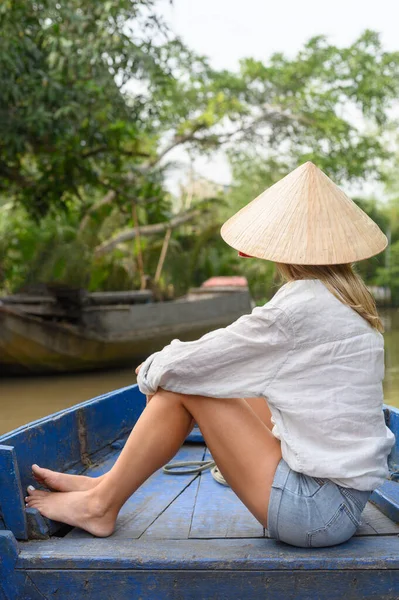 stock image back view of caucasian Female tourist in casual clothes and Vietnamese hat sitting with crossed legs and watching the nature on boat during boat trip in Mekong River Delta, Vietnam