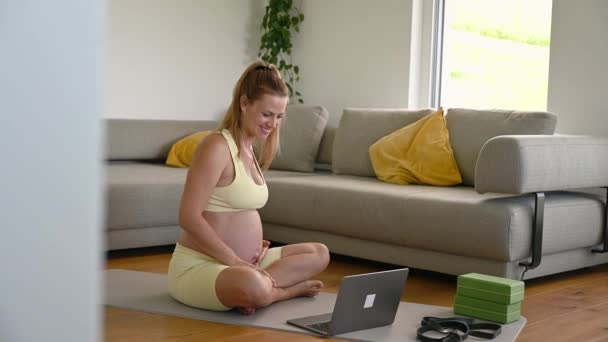 ソファーの近くのヨガマットに座っているスポーツウェアの若い妊婦の側面ビュー 朝のルーチン中にオンライン妊娠前のトレーニングをしている間 ノートパソコンの画面を見ます 高品質の4K映像 — ストック動画