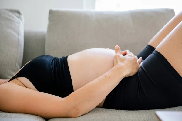 身着黑色运动服的怀孕妇女躺在沙发上 白天在家休息时摸着肚子的侧影 — 图库照片