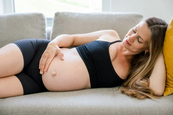 白天穿着黑色运动服的怀孕少女躺在沙发上摸着肚子在家休息的侧影 — 图库照片