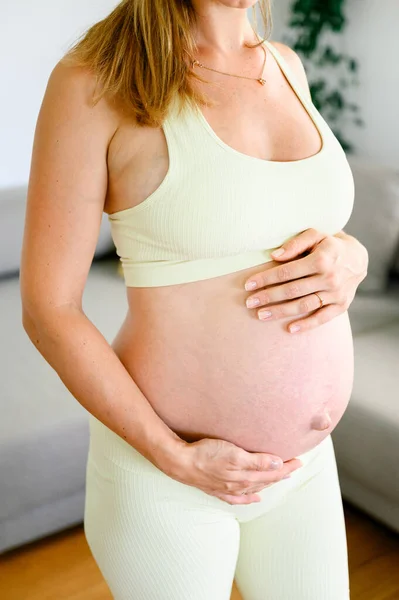 身着运动服的怀孕妇女站在客厅里 白天在家做完产前检查后双手靠在肚子上休息的罕见侧面景象 — 图库照片