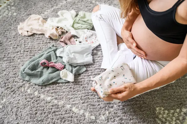 認識できない美しい妊婦のトップビュー 赤ちゃんのための準備をし 床に座って 腹に手を並べ 保育室で赤ちゃんの服を分類する — ストック写真
