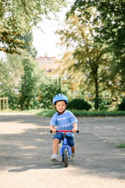 Güvenlik kaskı takmış şirin beyaz çocuk şehir parkındaki yolda bisiklet sürerken eğleniyor. Önce çocuk bisikleti. Çocuklar yaz sporları yapıyor.