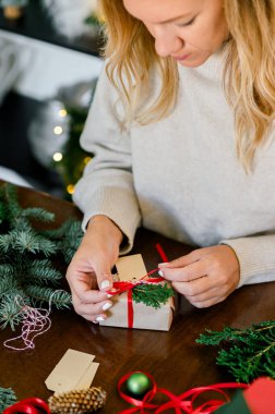 Kazaklı bir kadının Noel hediyelerini kağıt, makas, renkli kurdeleler kullanarak paketlemesi ve evdeki hediye kutusuna köknar koyması. Arka plan ışıklarında. Noel konsepti