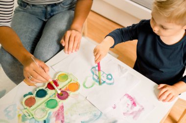 Sarışın, tatlı, sıradan görünümlü bir çocuk, anne evde oturuyor ve gündüz vakti resimleriyle resim çiziyor.