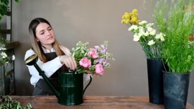 Ahşap tezgahın yanında duran önlüklü genç bayan çiçekçi ve çiçekçide iş günü sulama kutusunda buket düzenliyor.