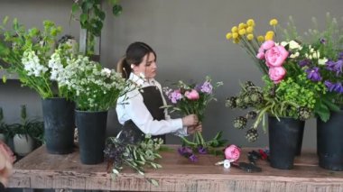 Çiçekçide çalışırken elinde bir buket aromatik çiçek ve yeşil yapraklı saksılarla masada duran genç bir bayan çiçekçi. iş görüntüleri