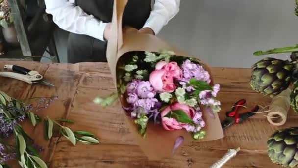 若い陽気な長い髪の女性の花屋エプロンは花屋で花束の香りの束を包んでいる間 木のテーブルに立っています ビジネスコンセプト 4K映像 — ストック動画