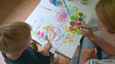 Anne ve oğlunun evde boya fırçaları ve suluboya boyalarla resim yapmalarının en üst görüntüsü. Genç bir kadın oturuyor ve oğluna resim çizmeyi öğretiyor. 4k video eğitim kavramı