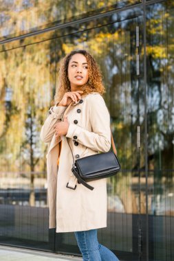 Moda bir ceket ve kot pantolonlu, gülümseyen ve kameraya bakan genç bir kadın.