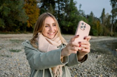 İnsanlar ve seyahat tutkusu. Zevkten dört köşe olmuş genç bir kadın selfie portresi yapıyor, güzel gölde boş zaman geçiriyor, yağmurluk giyiyor, sahilde tek başına duruyor. Eğlence ve gezi. Arkaplan ağaçları