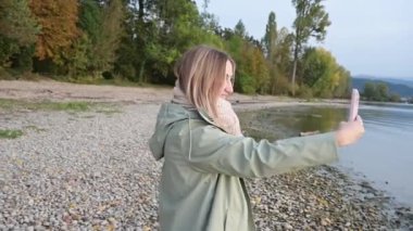 İnsanlar ve seyahat tutkusu. Zevkten dört köşe olmuş genç bir kadın selfie portresi yapıyor, güzel gölde boş zaman geçiriyor, yağmurluk giyiyor, sahilde tek başına duruyor. Eğlence ve gezi. Arkaplan ağaçları