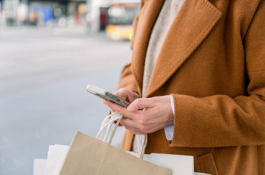 Şehrin caddesinde dikilen ve cep telefonuyla mesajlaşan, elinde alışveriş torbaları tutan, şık ceketli, isimsiz bir kadının yan görüntüsü.