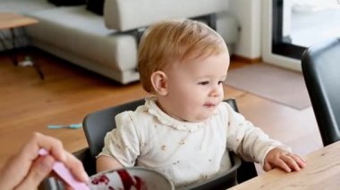 Mutfak masasında oturan ve bebeğini elma püresiyle besleyen mutlu bir kadın. Çocuk meyve püresi yerken kucağında bebeği tutan genç bir anne. Ebeveynlik, annelik kavramı 4k