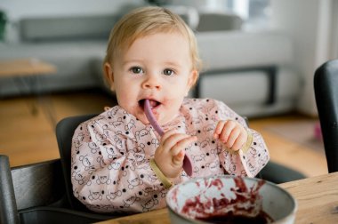 Gülümseyen bebek evde mutfaktaki masada tek başına kaşığı elinde tutuyor. Sevimli küçük çocuk kavanozdan meyve ya da sebze püresi alıyor. Bebeklik, çocukluk kavramı.