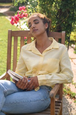Kıvırcık saçlı genç Afro-Amerikan kadın ahşap koltukta oturuyor, el ele kitap tutuyor, öğleden sonra dinleniyor ve dinleniyor kır evindeki bahçede arka plan çiçekleri pembe