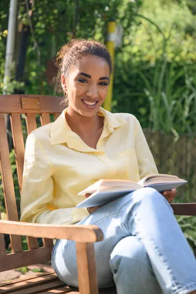 Young hipster black female in casual clothes, curly hair, holding book, reading looking at the camera, smiling in the garden at sunset. relaxing after work in the garden