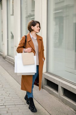 Moda montlu ve botlu genç bir kadın sokakta alışveriş çantasıyla dikilip dükkanın camına bakıyor, gündüz vakti şehirde yürüyor.