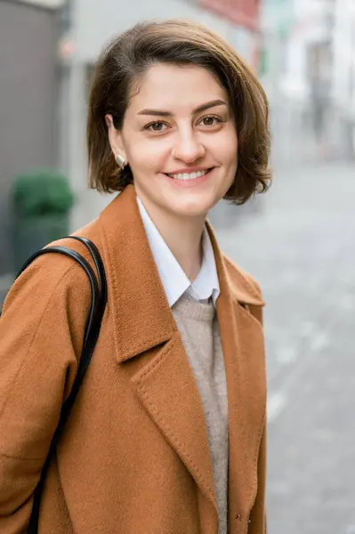 积极的年轻女性 棕色短发 戴着耳环 穿着雅致的外套 微笑着 站在城市街道上看着相机 — 图库照片