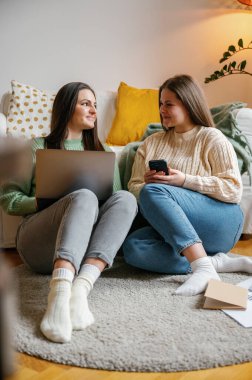 İki genç kadın, en iyi arkadaşlar rahat bir odada oturmuş dizüstü bilgisayar ve akıllı telefondan tercihlerini karşılaştırıyorlar muhtemelen internetten alışveriş yapıyorlar, rahat bir oturma odasında.