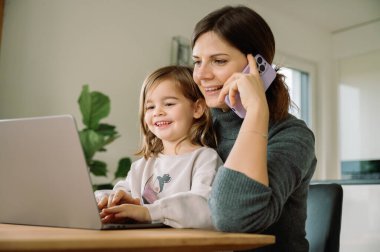 Rahat bir şekilde gülümseyen anne rahat bir ev ortamında dizüstü bilgisayarla kızını meşgul ederken aynı anda birden fazla iş yapıyor. iş hayatı aile kavramını dengeler