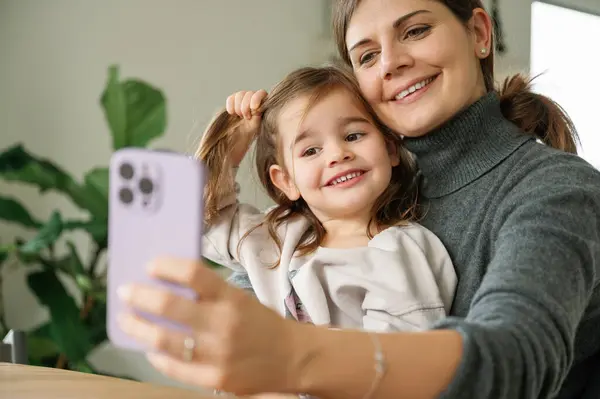 Cheerful mom and little daughter laughing using smartphone app at home sitting at table. Happy positive family parent having fun while making funny selfies, enjoying relaxing with little girl