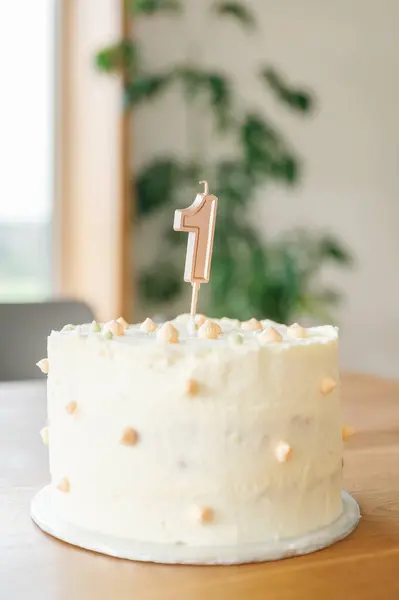 a first birthday cake with frosting and minimalistic decoration with a candle of number one on the table, in background living room. childhood concept