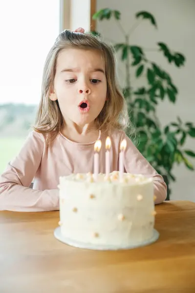 3歳の少女は テーブルに座っている明るいピンクのトップで ホームのお祝いで誕生日のケーキにろうそくを吹き飛ばします — ストック写真