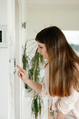 Gündelik giysiler içinde genç bir esmer kadının modern bir ev içinde dijital termostat kurarken yan görüntüsü.