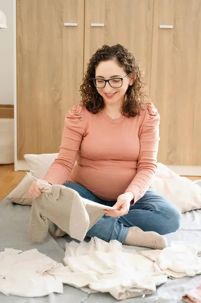 保育園の床に座っている幸せな妊婦の垂直写真は 昼間に赤ちゃんの服を折ります 妊娠と家族の概念 — ストック写真