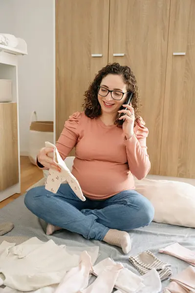 自宅で赤ちゃんの服を見ながら携帯電話を使用している妊婦の垂直写真 赤ちゃんの服を折りたたみ 出産のためのバッグを準備 家族の概念 — ストック写真