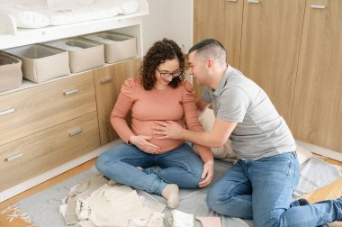 Müstakbel iki ebeveyn nedensel giysiler içinde yerde oturmuş bebek odasında birlikte mutlu bir şekilde bebek bekliyorlar. Erkeğin kadını kucaklaması, birlikte gülümsemesi, doğum konsepti.