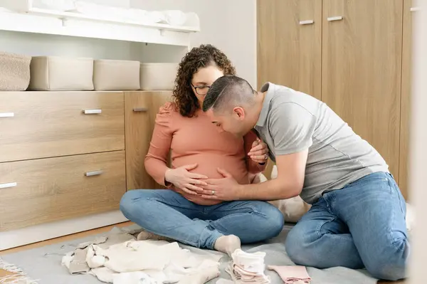 男人摸着他怀孕的妻子的肚子 在她肚子里和女婴说话 坐在婴儿房的地板上 背景婴儿服装 怀孕和人的概念的前景 — 图库照片