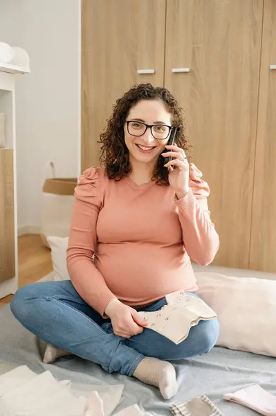 孕妇与电话交谈 在卧室里整理婴儿衣服 期待婴儿 怀孕和家庭观念的垂直照片 — 图库照片