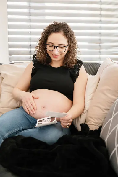 一个快乐的孕妇俯视着她的腹部 抱着超声波坐在沙发上 穿着休闲装和眼镜的垂直照片 怀孕概念 — 图库照片