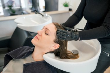 Bir kadın saçını kuaförde yıkatıyor. Siyah gömlek ve eldiven giyen kuaför. Müşteri gülümsüyor ve rahatlamış görünüyor. Salonda iki lavabo var. güzellik konsepti