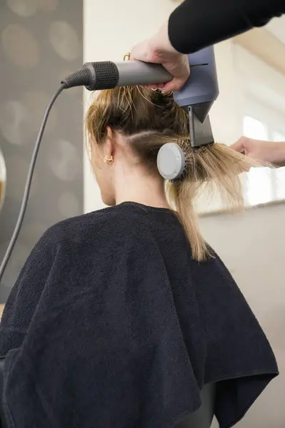 back view of unrecognizable woman is getting her hair blow dried by a stylist. The stylist is using a round brush to style the womans hair