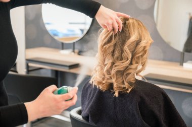 Kuaförü saç spreyiyle saçını yaptırırken, kuaföründe oturan ve aynaya bakan tanınmayacak kadar güzel genç bir kadının arka plan görüntüsü.