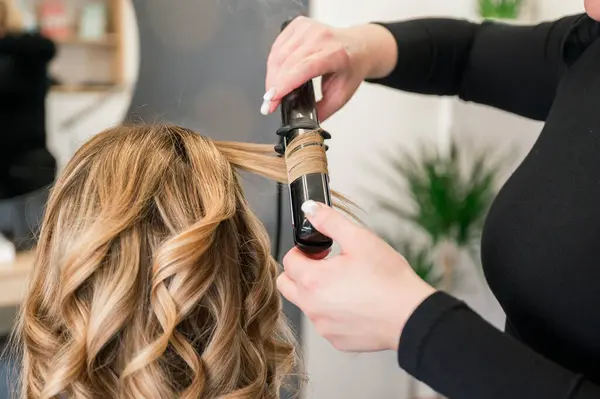 unrecognizable Hairstylist is styling a womans hair with a curling iron. The woman is wearing a black shirt and is standing in front of a potted wall, background beauty salon