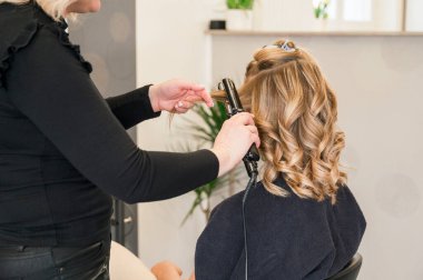 Tanımlanamayan bir kadın saçını bir kuaföre yaptırıyor. Kuaför, arka plandaki kadın saçını, güzellik salonunu, iş ve güzellik konseptini düzleştirmek için ütü kullanıyor.